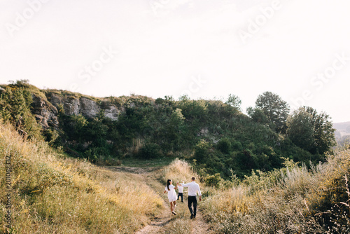 Walk beautiful young family in white clothes with a young son blond in mountainous areas with tall grass at sunset. Mother keeps son in her arms, hugging. family - this is happiness © Oleksandr