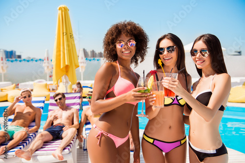 Three hot international ladies best friends are having drinks at the pool party, boyfriends are loking at them and smiling, lying on beach chairs, sunbathing, at roof top summer chill club
