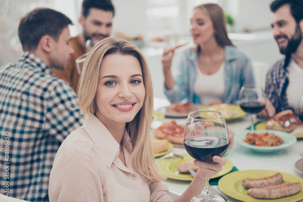 Charming, stylish, blonde girl sitting at the table with her best friends celebrating birthday looking at camera holding glass with red wine guests, visitors enjoying delicious, tasty, meals, dishes