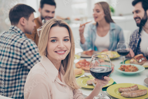 Charming  stylish  blonde girl sitting at the table with her best friends celebrating birthday looking at camera holding glass with red wine guests  visitors enjoying delicious  tasty  meals  dishes
