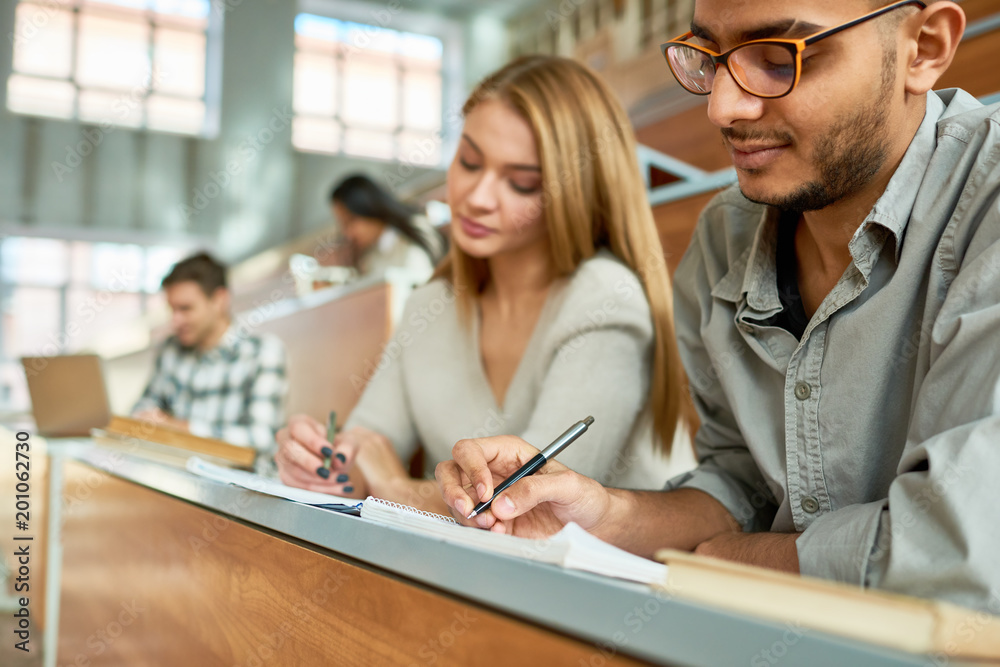 Multi-ethnic group of students sitting at desk in lecture hall of modern college and writing, focus on young Middle-Eastern man wearing glasses, copy space Multi-ethnic group of students sitting at desk in lecture hall of modern college and writing, focus on young Middle-Eastern man wearing glasses, copy space