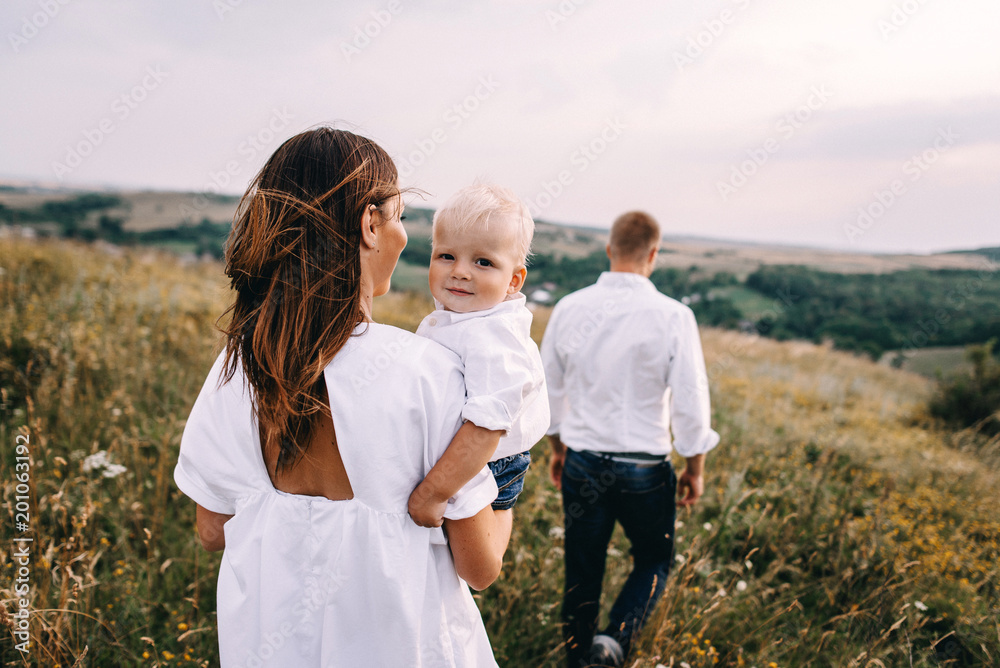 Walk of beautiful young family in white clothes with a young son blond in mountainous areas with tall grass at sunset. Dads carries his son on his shoulders. family - this is happiness