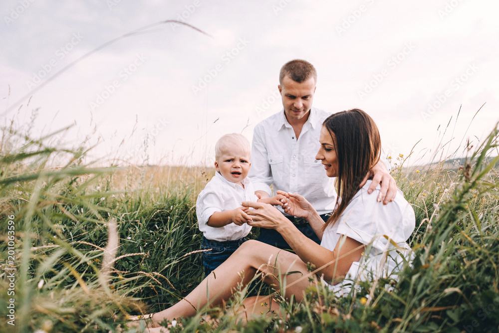 Walk beautiful young family in white clothes with a young son blond in mountainous areas with tall grass at sunset. Mother keeps son in his arms, hugging. family - this is happiness