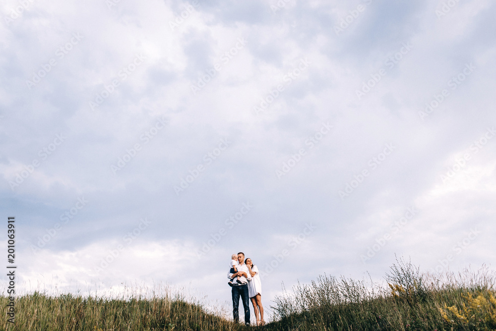 Walk of beautiful young family in white clothes with a young son blond in mountainous areas with tall grass at sunset. Family watching the sunset on the background of the beautiful sky Walk of beautiful young family in white clothes with a young son blond in mountainous areas with tall grass at sunset. Family watching the sunset on the background of the beautiful sky