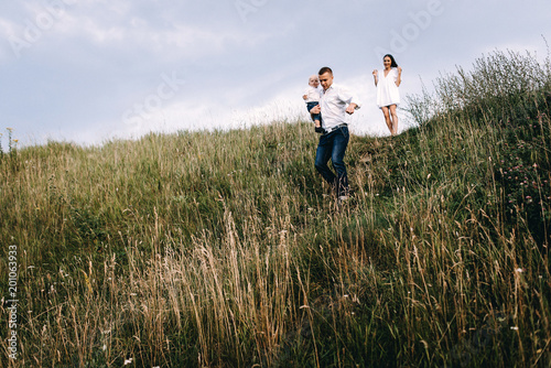 Walk of beautiful young family in white clothes with a young son blond in mountainous areas with tall grass at sunset. Family watching the sunset on the background of the beautiful sky © Oleksandr