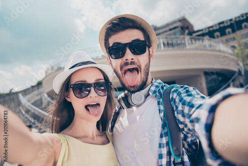 Foolish crazy comic funny couple in caps having journey trip meeting outdoors shooting self portrait on front camera showing tongue out, beautiful and handsome bloggers having video-call with friends