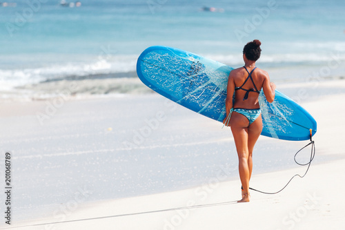 Beautiful pacific islander melanesian afro-american surfer girl in blue bikini carrying surfboard to the sea at Padang Padang beach, Bali, Indonesia photo