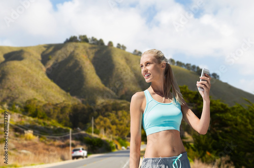 fitness, sport and technology concept - smiling young woman with smartphone exercising over big sur hills and road background in california