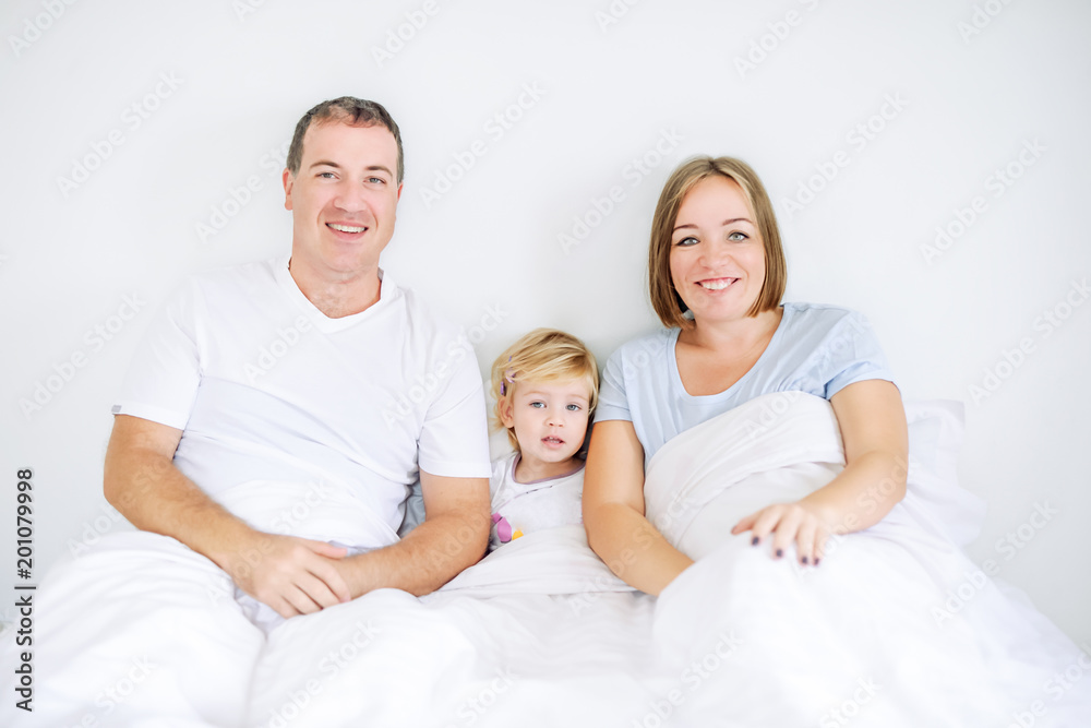 Portraits of happy and loving family in sleepwear looking at camera in the morning time. Cheerful parents having fun with baby daughter on the bed. Soft selective focus. Space for text.