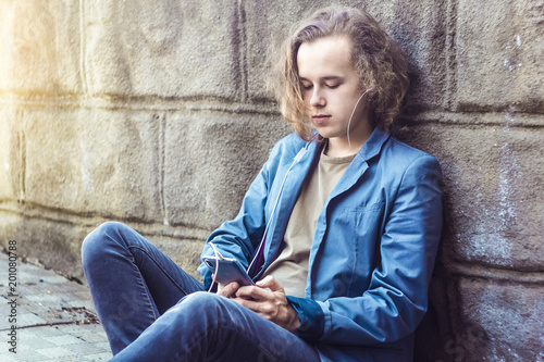 A teenager boy  listening to music and using a telephone in an urban setting. A handsome young man sitting on the sidewalk writing a text message.   © Oleksandr
