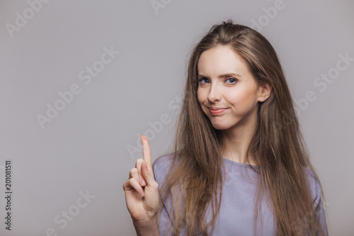 Horizontal portrait of pleasant looking brunette female model with dark hair, keeps fore finger raised upwards, shows something, poses against grey background with blank copy space for advertisment