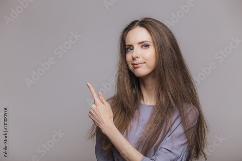 Shot of serious pleasant looking brunette female with blue eyes, dressed casually, points with index finger at blank copy space, isolated over grey background. Woman advertizes new product indoor