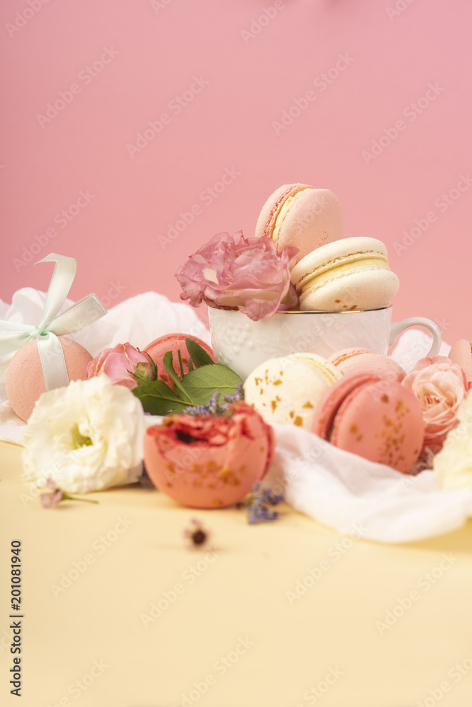 Pink and white macaroons cakes with big and small flower buds are decoratively laid on a white fabric and a cup on a yellow background Pink and white macaroons cakes with big and small flower buds are decoratively laid on a white fabric and a cup on a yellow background