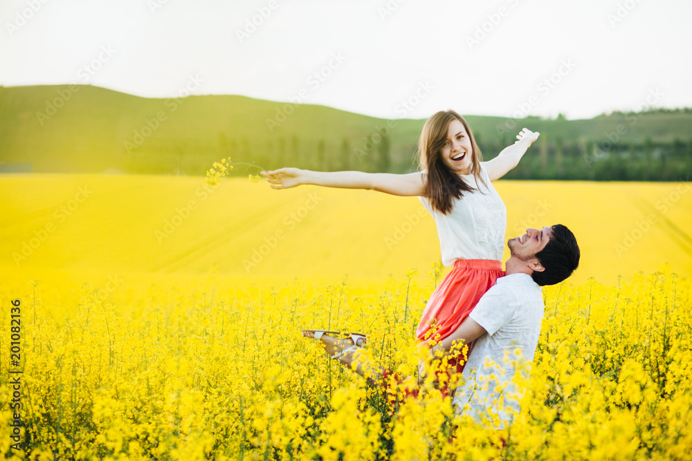 Romantic man raises his girlfriend, enjoy togetherness at beautiful yellow field, pose outdoor, have fun, smile joyfully. Couple in love spend summer holidays at nature. People and harmony concept Romantic man raises his girlfriend, enjoy togetherness at beautiful yellow field, pose outdoor, have fun, smile joyfully. Couple in love spend summer holidays at nature. People and harmony concept