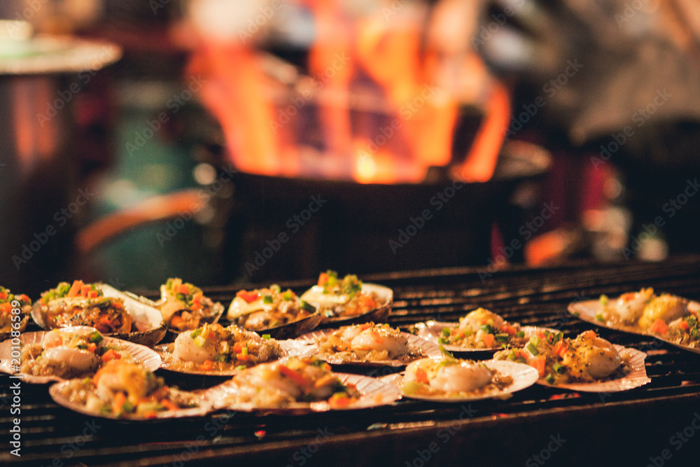 Grilled scallops sold at a street market stall with a pan covered with flames on the background. Chinatown, Bangkok, Thailand