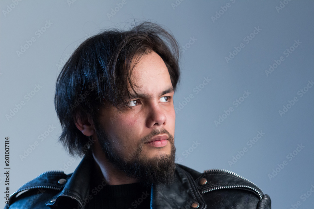 A stern gaze of a guy in a black leather jacket with a beard and long hair in the style of rock'n'roll. Male portrait in studio looking away. A stern gaze of a guy in a black leather jacket with a beard and long hair in the style of rock'n'roll. Male portrait in studio looking away.