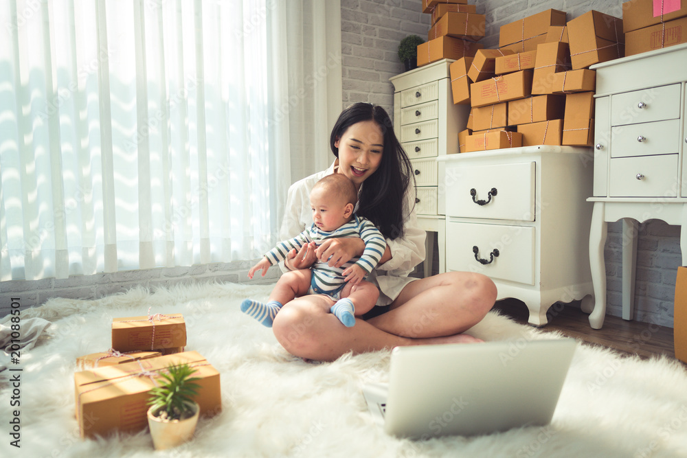 Businesswoman with baby boy working at home using laptop and preparing package product on background. SME entrepreneur or freelance life style concept. - obrazy, fototapety, plakaty 