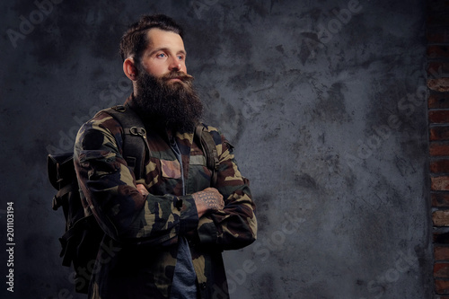 A handsome bearded traveler, dressed in a trendy camouflaged jacket, with a tattoed arms, standing in a studio, isolated on a dark background.