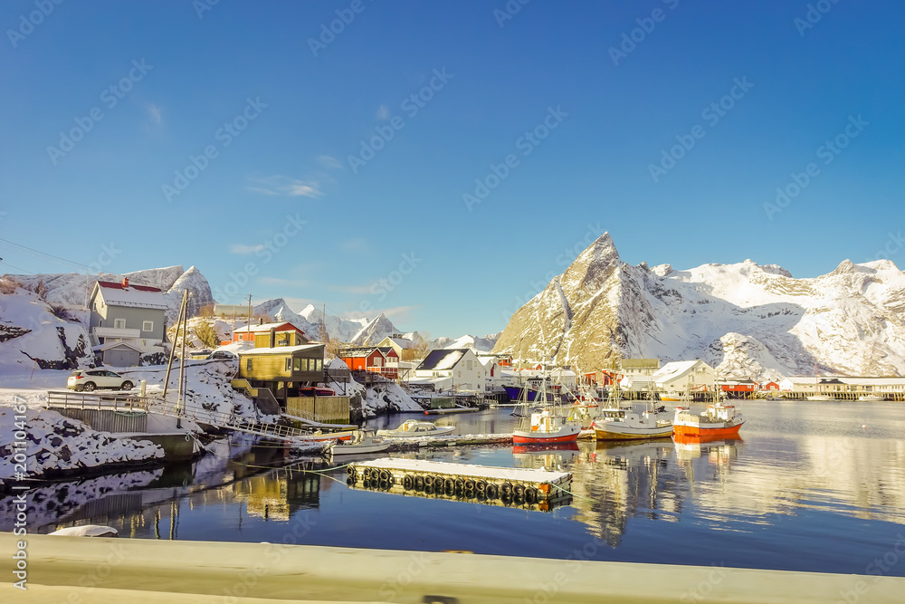 Above view of some wooden buildings in the bay with boats in the shore in Lofoten Islands surrounded with snowy mountains and colorful winter season