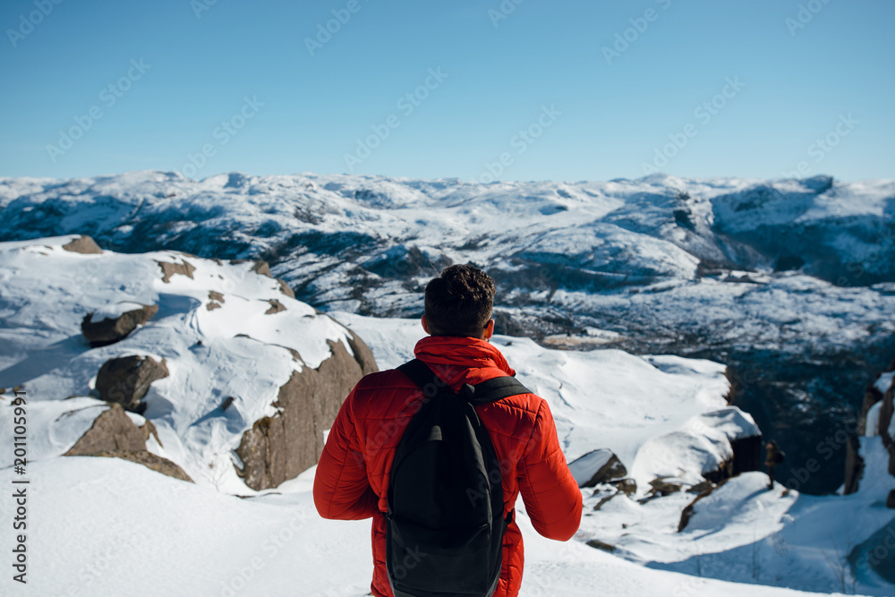 Rear view of a traveler man in red jacket and black backpack looking at the beautiful snowy mountain landscape near the Pulpit Rock, Preikestolen, Lysefjord, Norway.