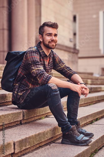 Beautiful male with stylish haircut and beard, wearing a fleece shirt and jeans, sitting on steps against an old building.