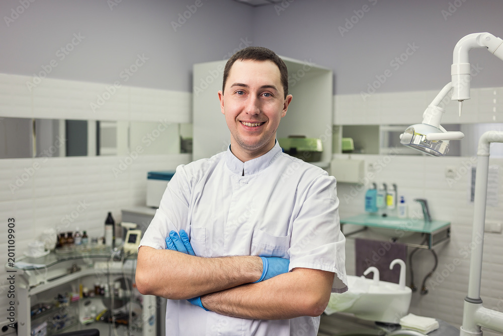 Obraz premium Male dentist standing with his hands crossed over medical office background. Healthcare, profession, stomatology and medicine concept