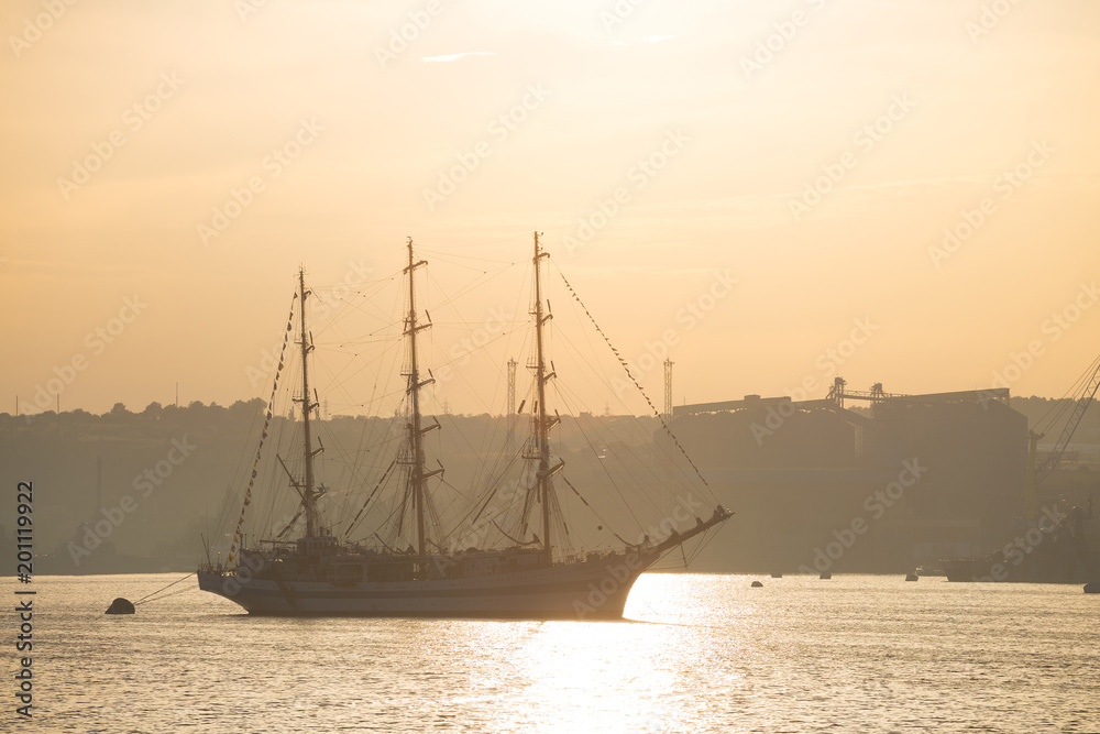 Three-masted sailing ship anchored in the port of Sevastopol at sunrise. Silhouettes of cargo port facilities and a warship on the background.