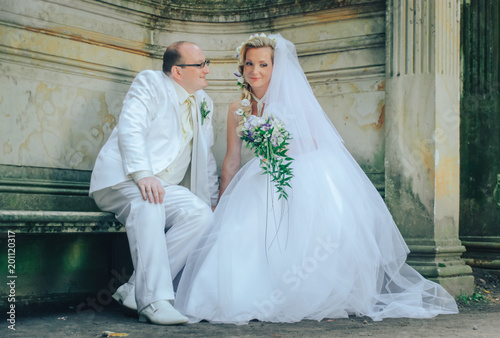 Wedding ceremony in nature.  Lovers newlyweds sitting on a bench in the Park, the groom in a white tuxedo, looking at his young wife.