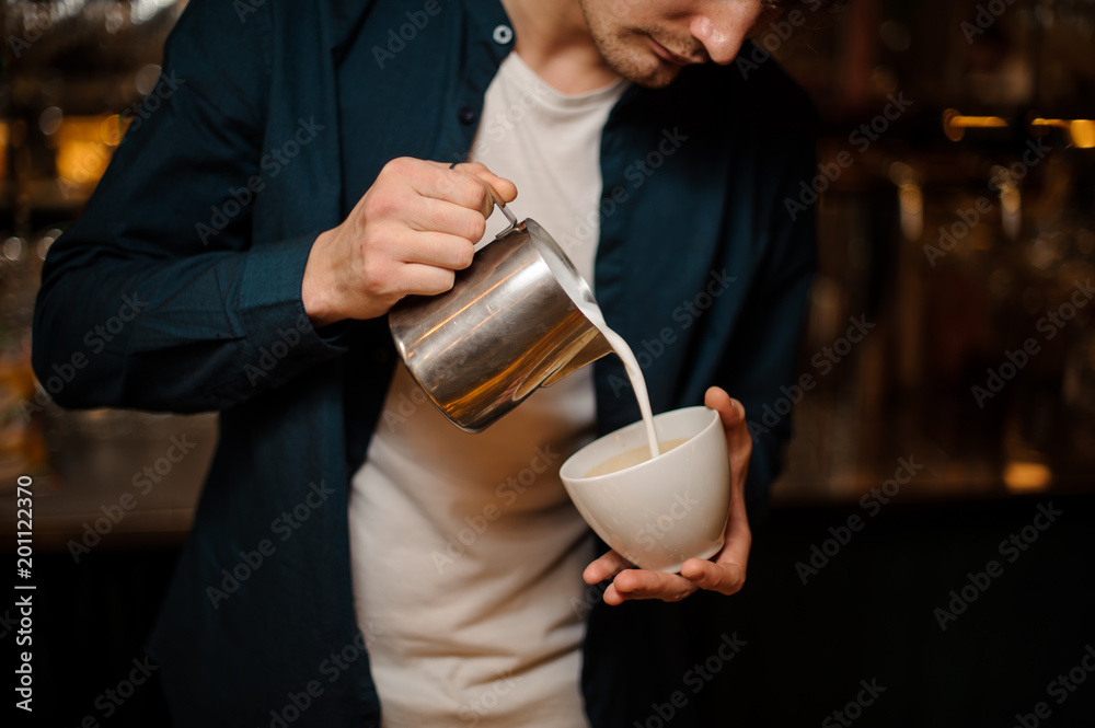 Brunet bartender pouring a milk into the white ceramic cup of coffee Brunet bartender pouring a milk into the white ceramic cup of coffee