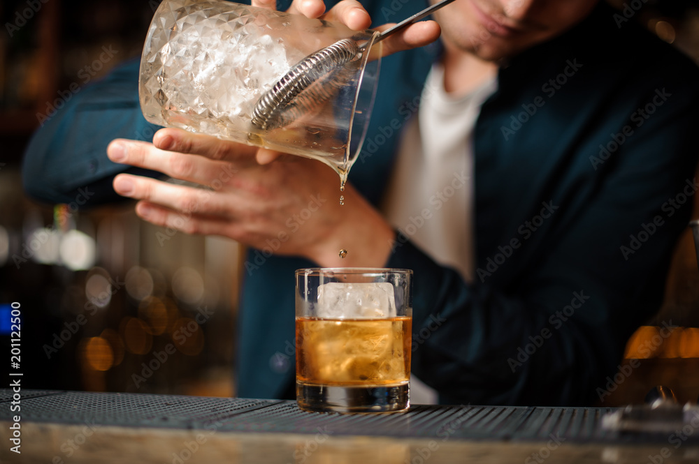 Brunet bartender pouring an alcoholic drink into a glass with an ice cube