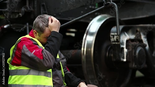 Stressed railroadman on railway track, railway worker sits on railway line near big metal wheel. Tired railwayman in red hard hat sits on rail near freight wagon after hard work day photo