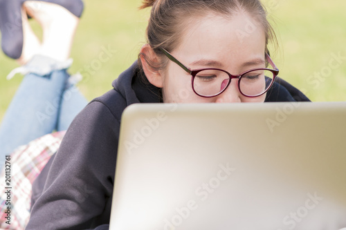 Young Asian woman wearing glasses lying at the grassy meadow and studying on her laptop.