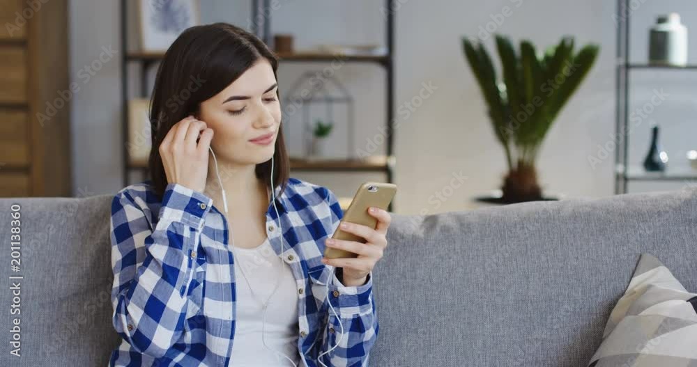 Young beautiful woman in the motley shirt taking on headphones and listening to the music on the smartphone in the cozy home. Indoor Young beautiful woman in the motley shirt taking on headphones and listening to the music on the smartphone in the cozy home. Indoor