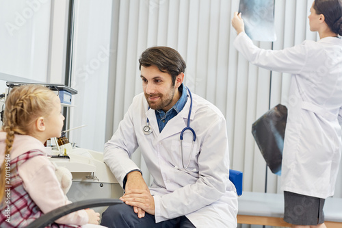 Male doctor talking with little patient while his colleague working in the background