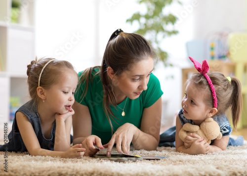 Young mother reading a book to her kids daughters. Children and mom lying on rug in cozy living room.