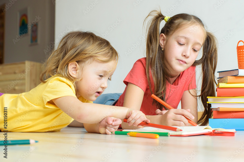 Two girls of eight and two years draw colored pencils, play on a floor. Children's creativity. Homescholing. Two children draw. Two girls of eight and two years draw colored pencils, play on a floor. Children's creativity. Homescholing. Two children draw.