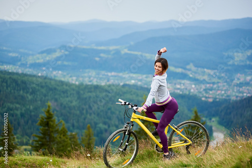 Beautiful female cyclist posing on yellow bicycle in the mountains on summer evening. Mountains, forests and small city on the blurred background. Outdoor sport activity. Copy space