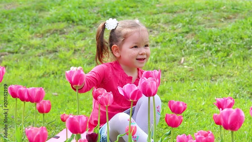Portrait of funny girl enjoying nature in urban park. Cute little child playing with fresh pink flowers while sitting on green lawn outdoors on spring sunny day. Portrait of funny girl enjoying nature in urban park. Cute little child playing with fresh pink flowers while sitting on green lawn outdoors on spring sunny day.