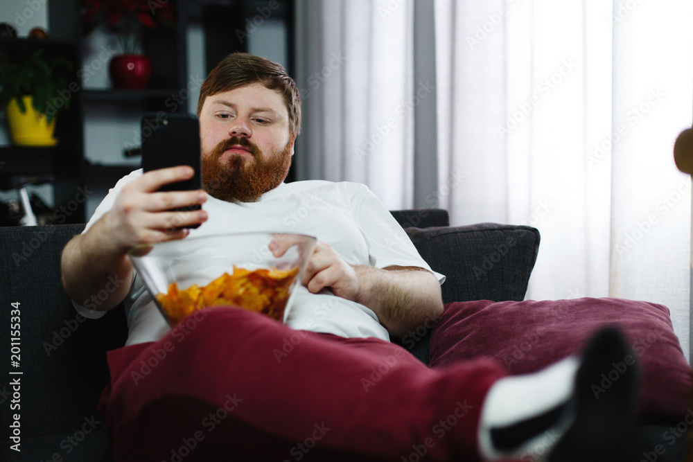 Pretty fat man smiles checking his smartphone while he sits on the sofa and eats