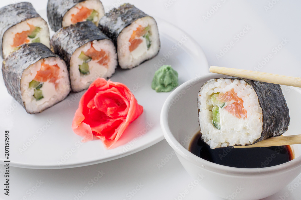 Sushi roll in chopsticks in soy sauce, closeup. Plate with roll japanes seafood sushi with ginger and wasabi on background. Sushi roll in chopsticks in soy sauce, closeup. Plate with roll japanes seafood sushi with ginger and wasabi on background.