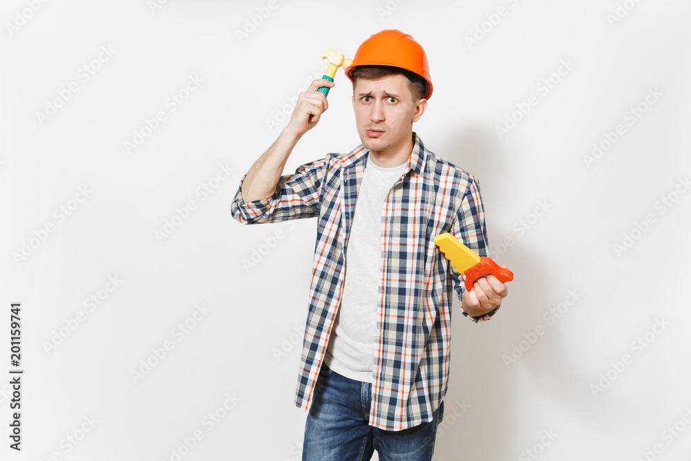 Young concerned man in protective orange hardhat holding toy saw and beating himself on head with toy hammer isolated on white background. Tools for renovation apartment room. Repair home concept. Young concerned man in protective orange hardhat holding toy saw and beating himself on head with toy hammer isolated on white background. Tools for renovation apartment room. Repair home concept.