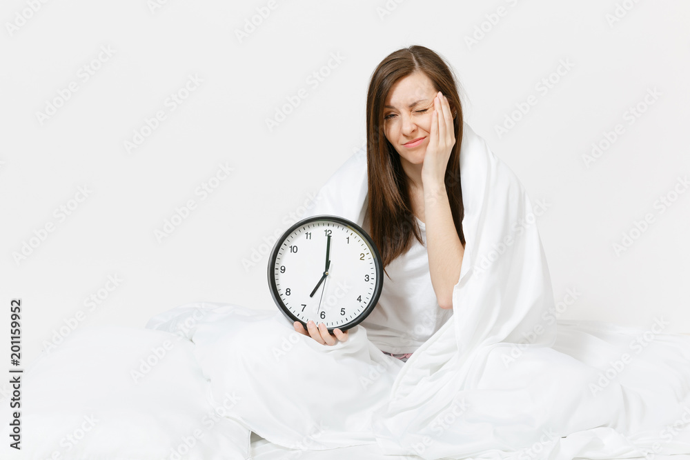 Young woman sitting in bed with round clock, white sheet, pillow, wrapping in blanket on white background. Beauty female wake up early in morning, spending time in room. Rest, relax, good mood concept Young woman sitting in bed with round clock, white sheet, pillow, wrapping in blanket on white background. Beauty female wake up early in morning, spending time in room. Rest, relax, good mood concept
