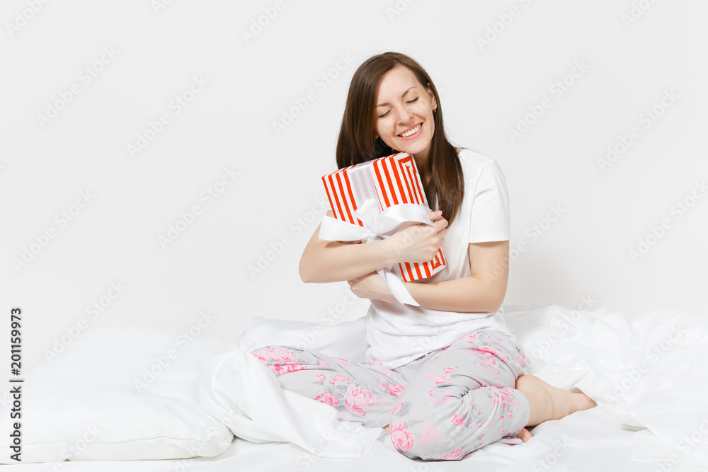 Young brunette woman sitting in bed with red striped gift box, white sheet, pillow, wrapping in blanket isolated on white background. Happy female spending time in room. Rest relax good mood concept.