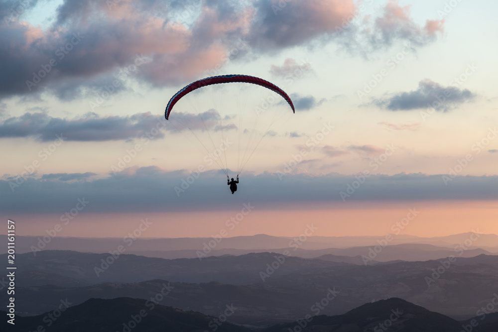 Beautiful shot of a paraglider silhouette flying over Monte Cucco (Umbria, Italy) with sunset on the background, with beautiful colors and dark tones Beautiful shot of a paraglider silhouette flying over Monte Cucco (Umbria, Italy) with sunset on the background, with beautiful colors and dark tones