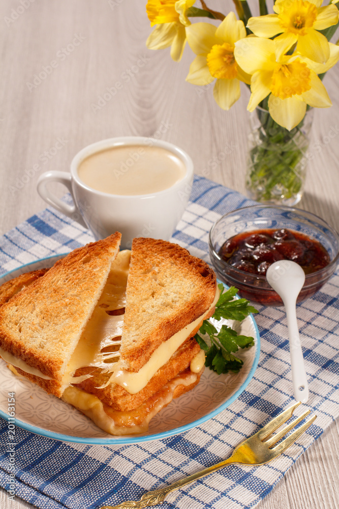 Toasted slices of bread with cheese and green parsley on white plate, fork, cup of coffee, bowl with strawberry jam, spoon and yellow daffodils on napkin Toasted slices of bread with cheese and green parsley on white plate, fork, cup of coffee, bowl with strawberry jam, spoon and yellow daffodils on napkin