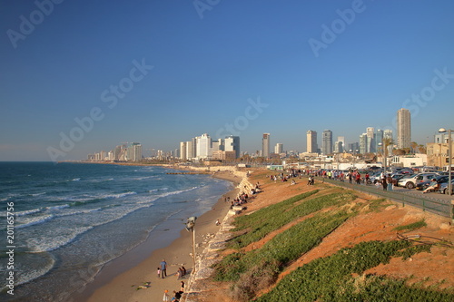 Tel Awiw, Izrael, widok na zatokę morza, nabrzeże z zielenią i umocnieniami, odpoczywających ludzi, ulicę z zaparkowanymi samochodami, w tle drapacze chmur, słonecznie