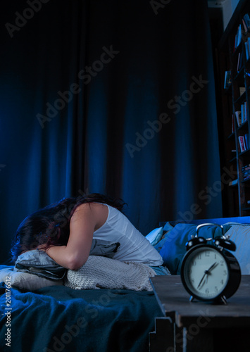 Photo of dissatisfied brunette with insomnia sitting on bed next to alarm clock