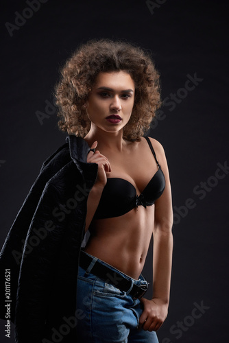 Sexy woman posing in black bra and blue jeans. keeping black jacket .Looking beatiful,confident, charismatic. Having pretty face features, many little curles and day make up. Studio black background.