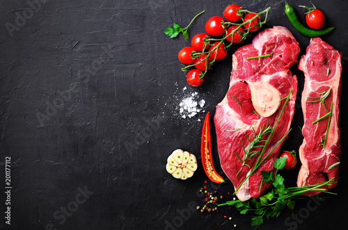 Raw meat steaks with cherry tomatoes, hot pepper, garlic, oil and herbs on dark stone, concrete background. Free space for your text.
