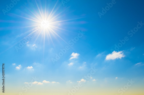 Sun with bright rays in the blue sky with white light clouds, at the horizon a yellowish shade, a gradient.